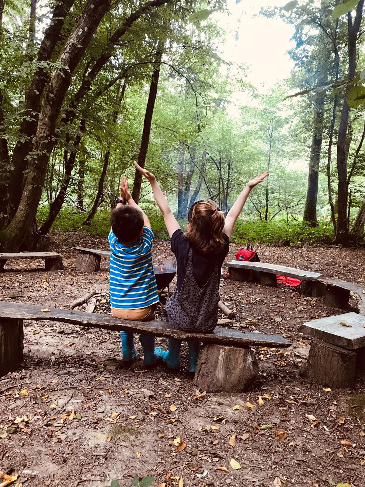 Two children sit around a fire circle in the woods with their arms raised up in celebration